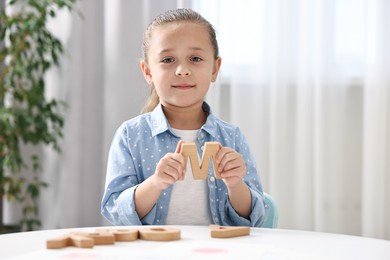 Photo of Learning alphabet. Little girl with wooden letter M at white table indoors