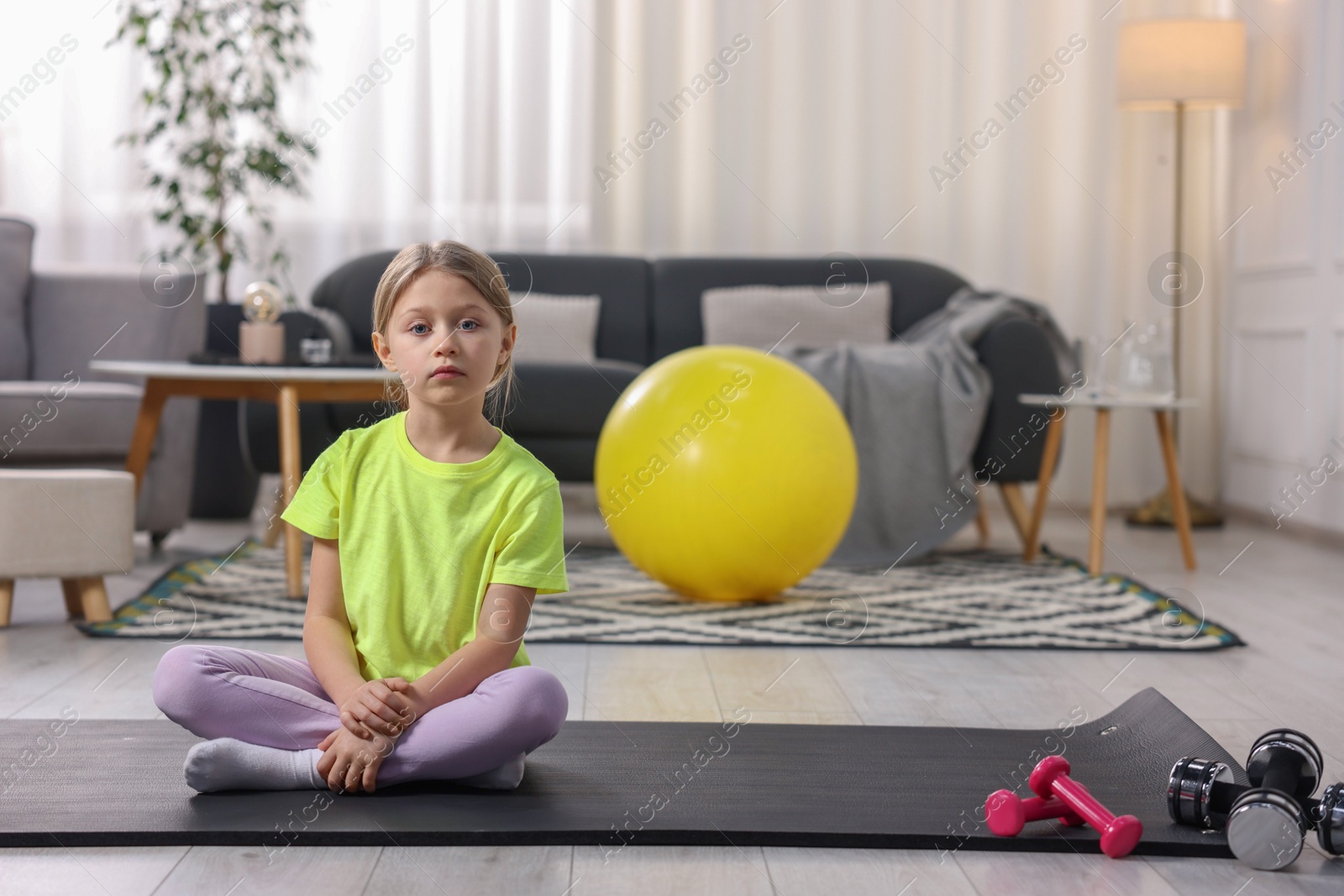 Photo of Cute little girl sitting on fitness mat indoors, space for text