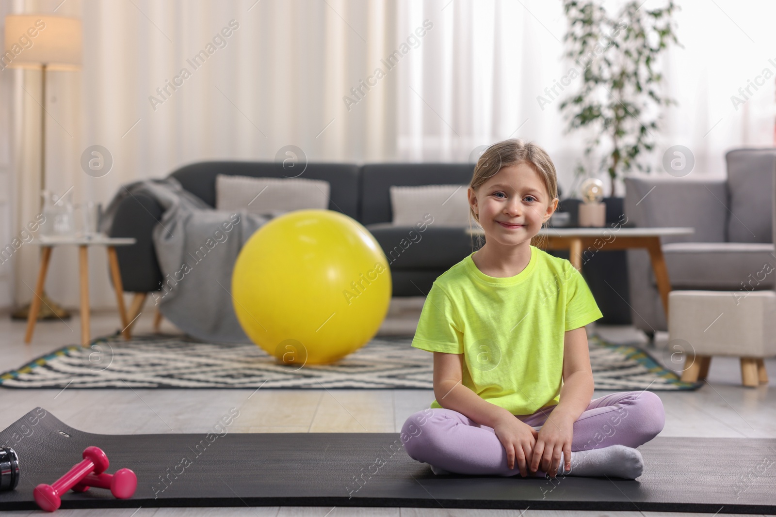 Photo of Cute little girl sitting on fitness mat indoors, space for text