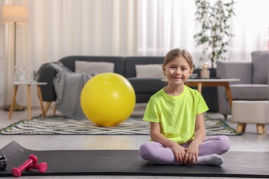 Cute little girl sitting on fitness mat indoors, space for text