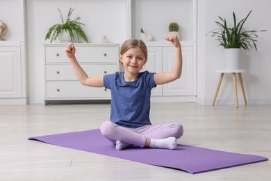 Photo of Cute little girl showing her biceps on fitness mat indoors