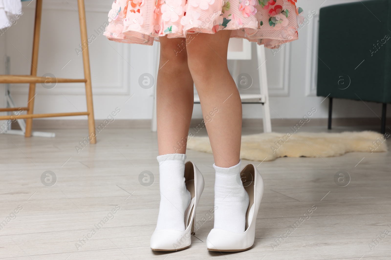 Photo of Little girl wearing oversized high heeled shoes indoors, closeup