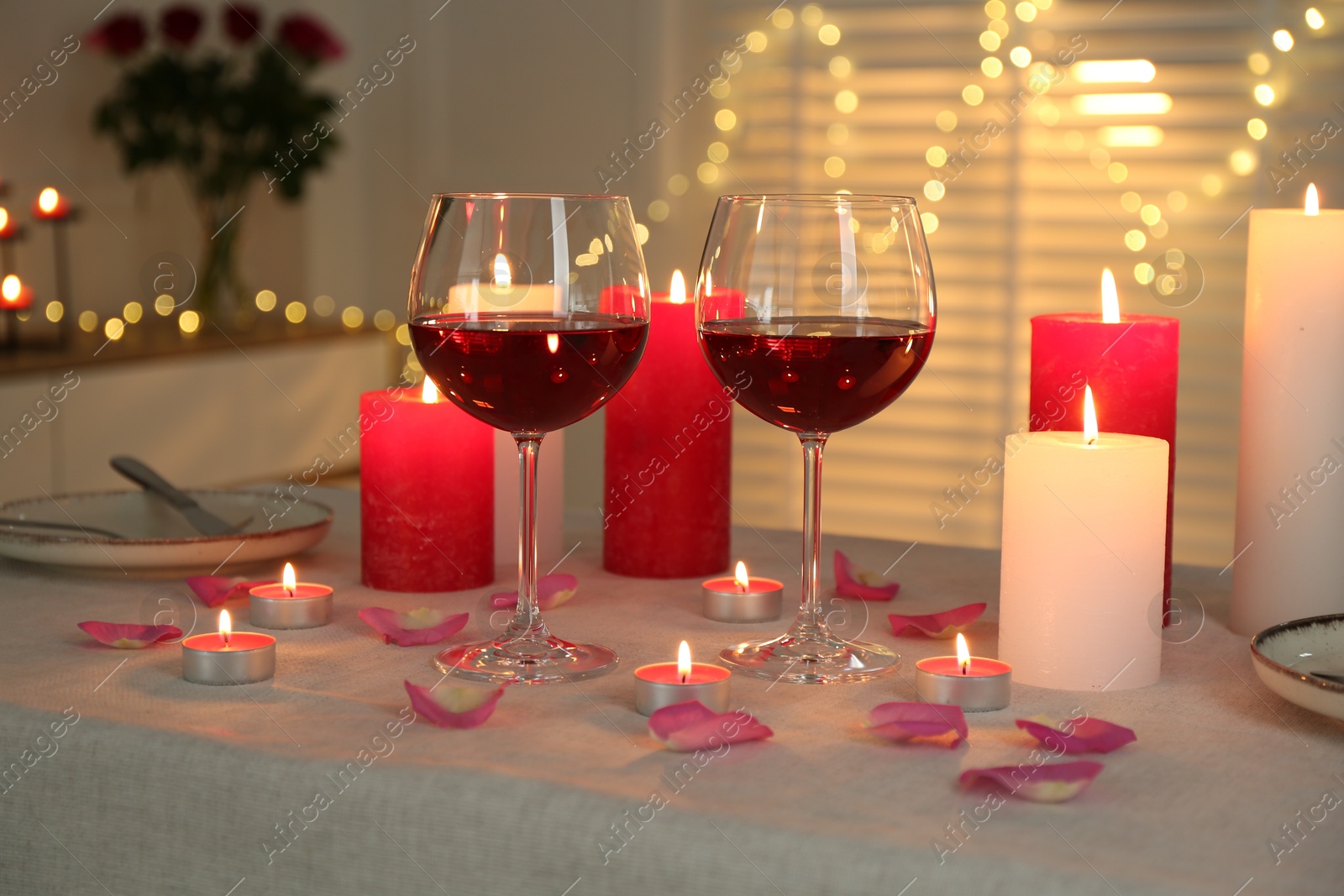 Photo of Romantic place setting with red wine, burning candles, rose and petals on table against blurred lights