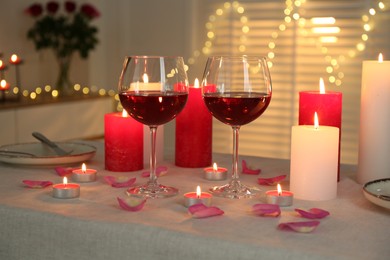 Romantic place setting with red wine, burning candles, rose and petals on table against blurred lights