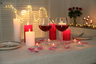 Romantic place setting with red wine, burning candles, rose and petals on table against blurred lights