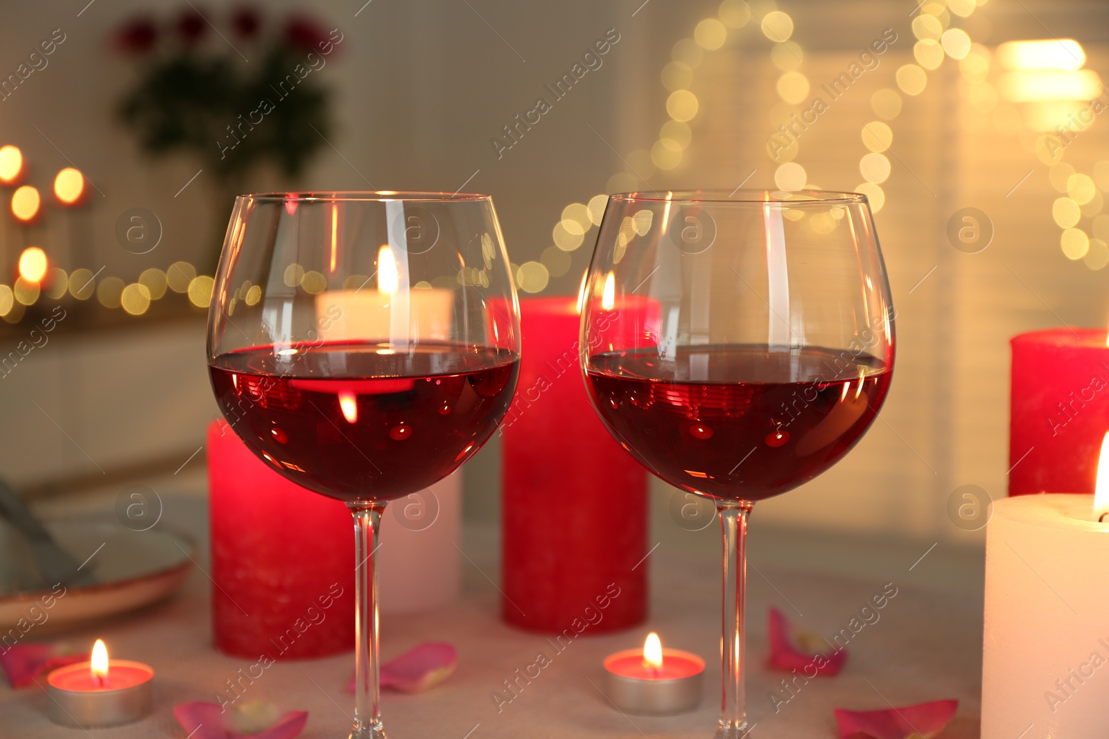 Photo of Romantic place setting with red wine, burning candles and petals on table against blurred lights, closeup