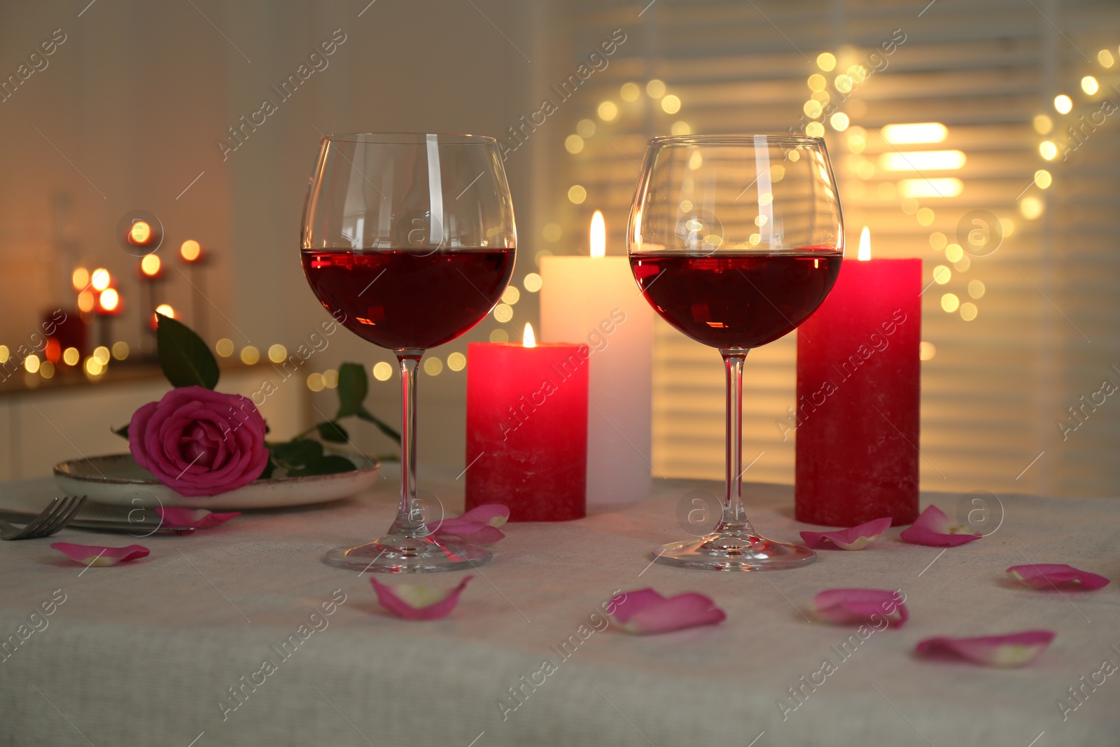 Photo of Romantic place setting with red wine, burning candles, rose and petals on table against blurred lights