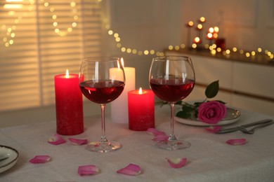 Romantic place setting with red wine, burning candles, rose and petals on table against blurred lights