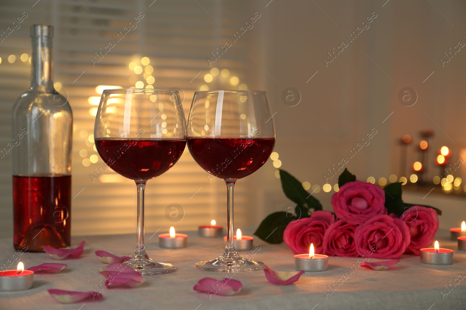 Photo of Romantic place setting with red wine, burning candles and roses on table against blurred lights