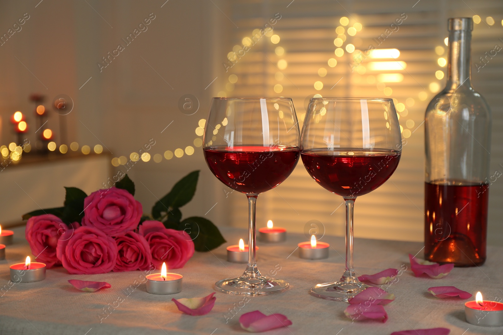 Photo of Romantic place setting with red wine, burning candles and roses on table against blurred lights