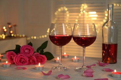 Photo of Romantic place setting with red wine, burning candles and roses on table against blurred lights