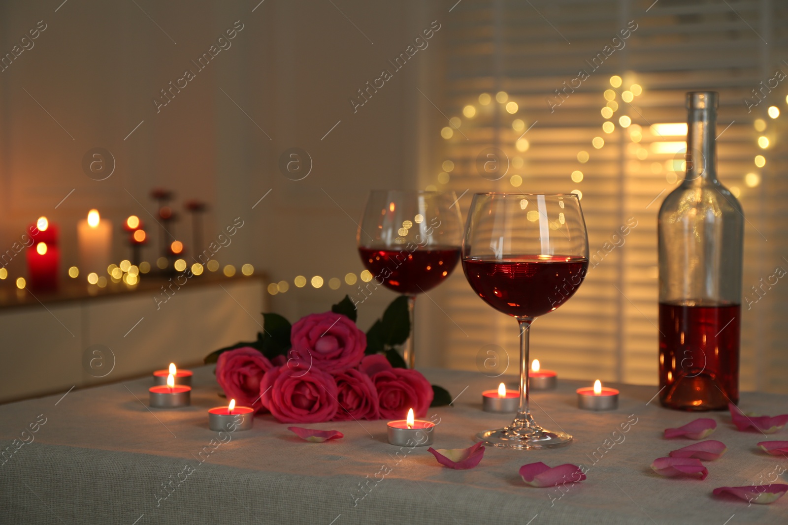 Photo of Romantic place setting with red wine, burning candles and roses on table against blurred lights