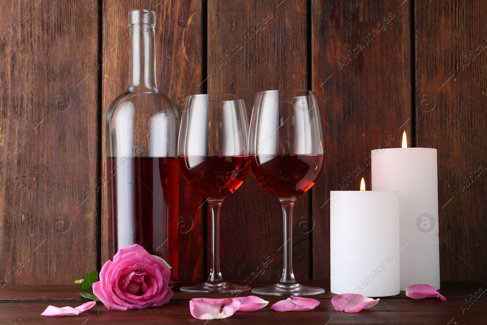 Photo of Glasses of red wine, bottle, burning candles and rose on wooden table