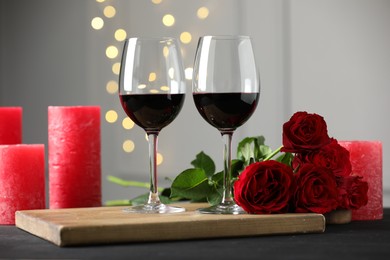 Glasses of red wine, candles and roses on gray table against blurred lights. Romantic dinner