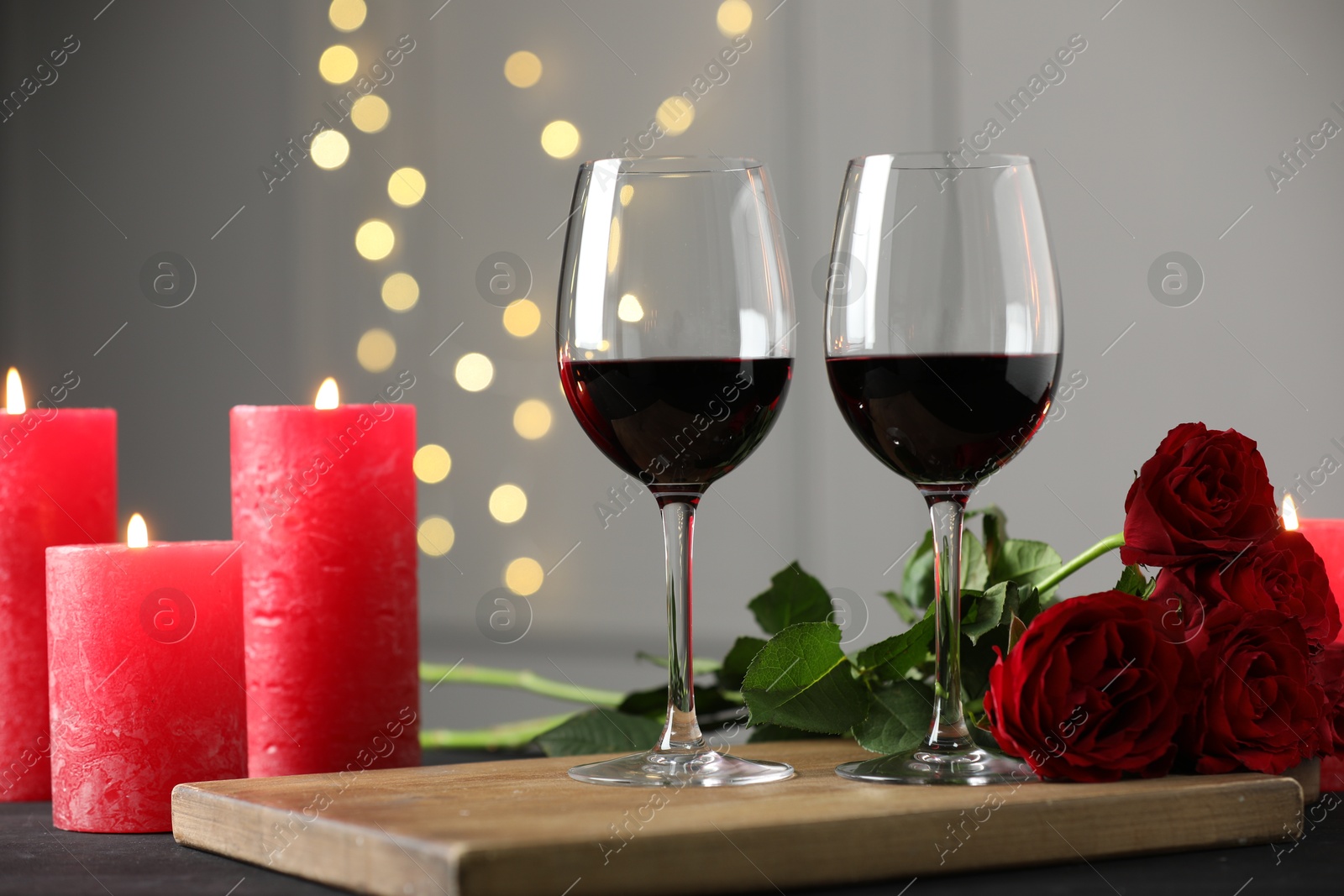 Photo of Glasses of red wine, burning candles and roses on table against gray background with blurred lights. Romantic dinner