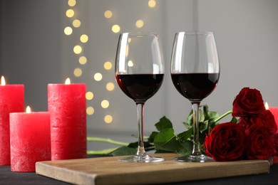 Photo of Glasses of red wine, burning candles and roses on table against gray background with blurred lights. Romantic dinner