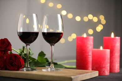 Photo of Glasses of red wine, burning candles and roses on gray table against blurred lights. Romantic dinner