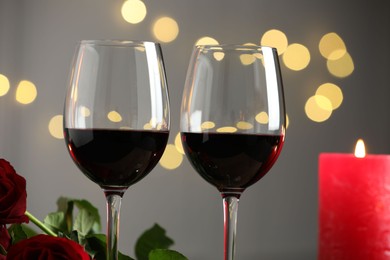 Glasses of red wine, burning candle and roses against blurred background, closeup. Romantic dinner