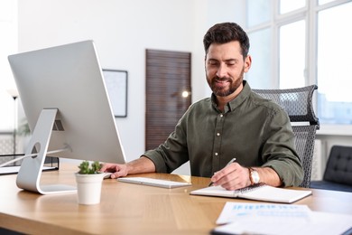 Photo of Man taking notes while working on computer at table in office