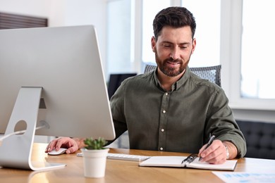 Photo of Man taking notes while working on computer at table in office