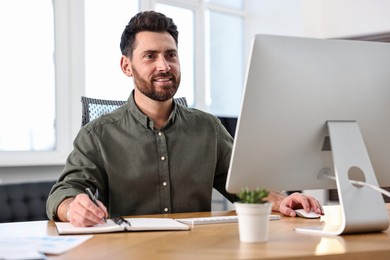 Photo of Man taking notes while working on computer at table in office