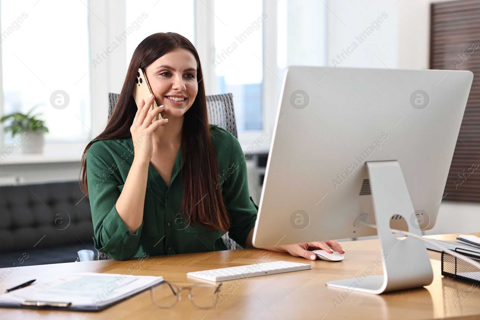 Photo of Woman talking on smartphone while working with computer at table in office