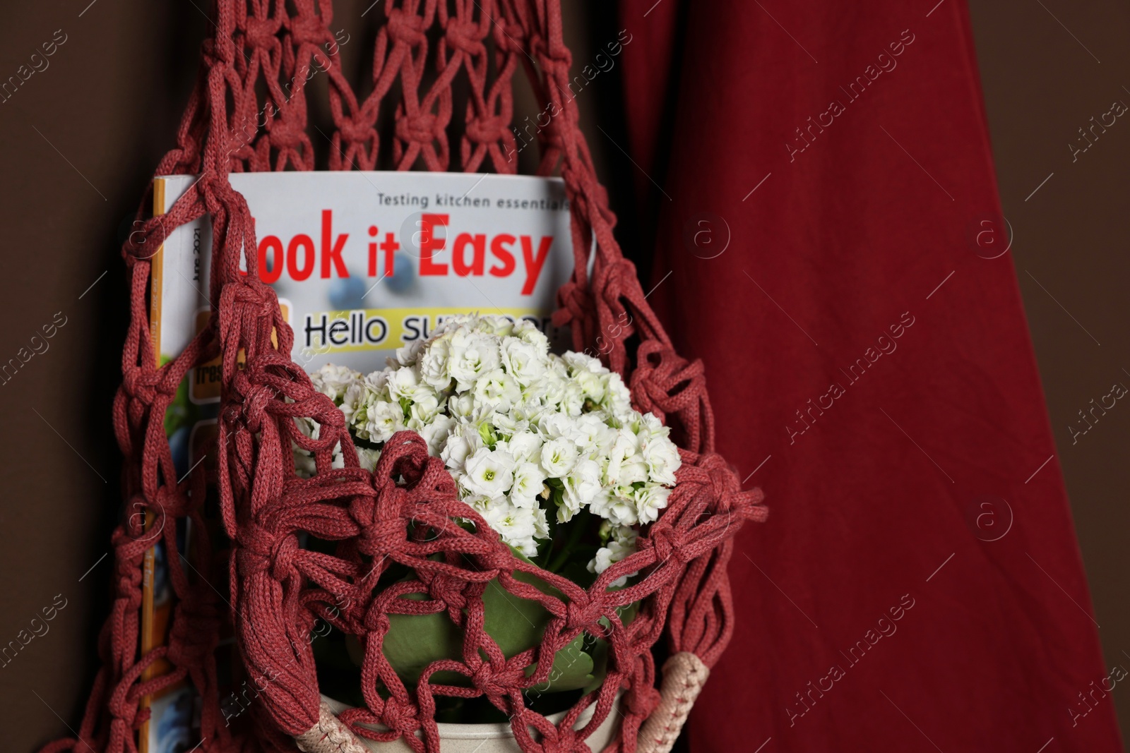 Photo of Handmade macrame shopping bag with magazine, flowers and shirt on brown wall, closeup