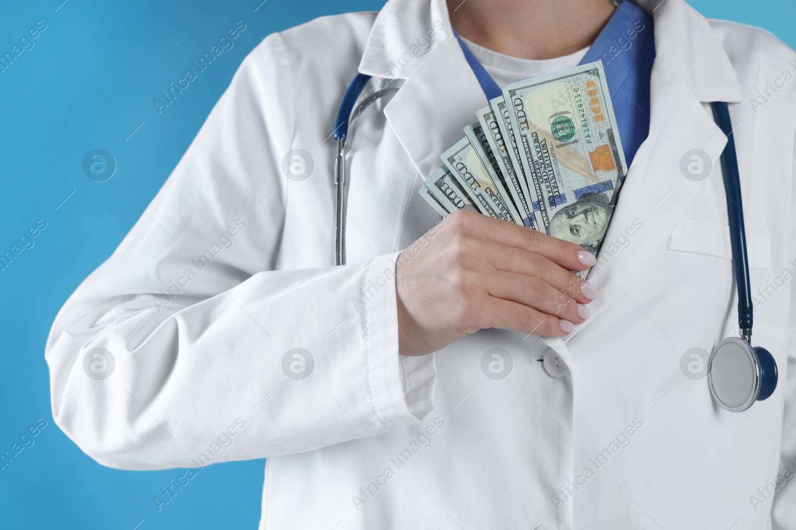 Photo of Corruption concept. Doctor putting dollar banknotes into her coat on light blue background, closeup