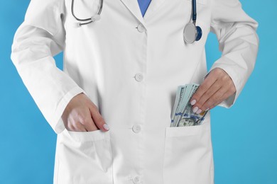 Photo of Corruption concept. Doctor putting dollar banknotes into her pocket on light blue background, closeup