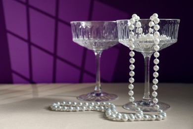 Photo of Elegant wine glasses and pearl jewelry on beige table against purple background, space for text