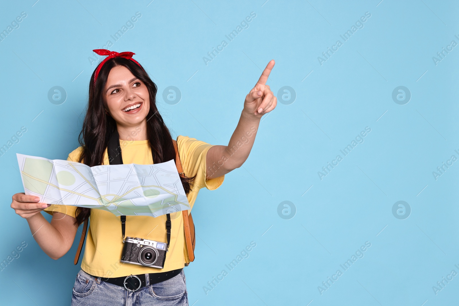 Photo of Young tourist in hat with camera and map pointing at something on light blue background, space for text