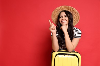 Photo of Young tourist in hat with suitcase pointing at something on red background, space for text