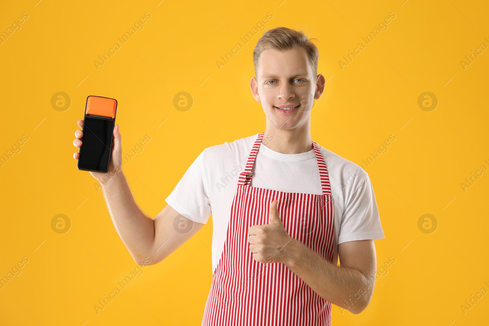 Photo of Happy young man in apron with payment terminal showing thumbs up on yellow background