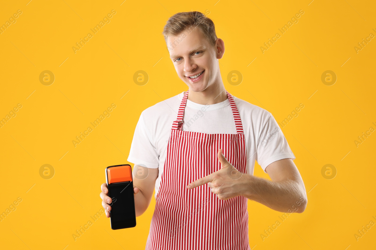 Photo of Happy young man in apron with payment terminal on yellow background