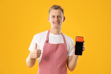 Happy young man in apron with payment terminal and debit card showing thumbs up on yellow background