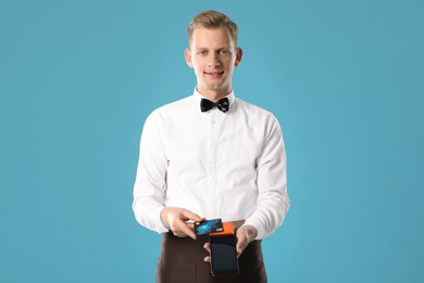 Happy waiter with payment terminal and debit card on light blue background