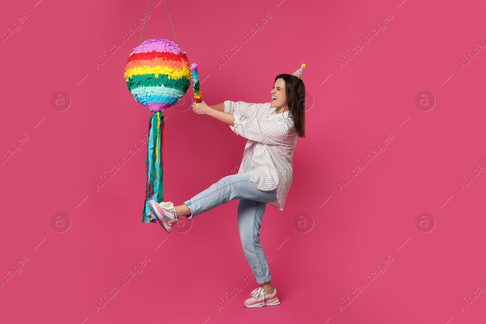 Photo of Happy woman hitting colorful pinata with stick on pink background