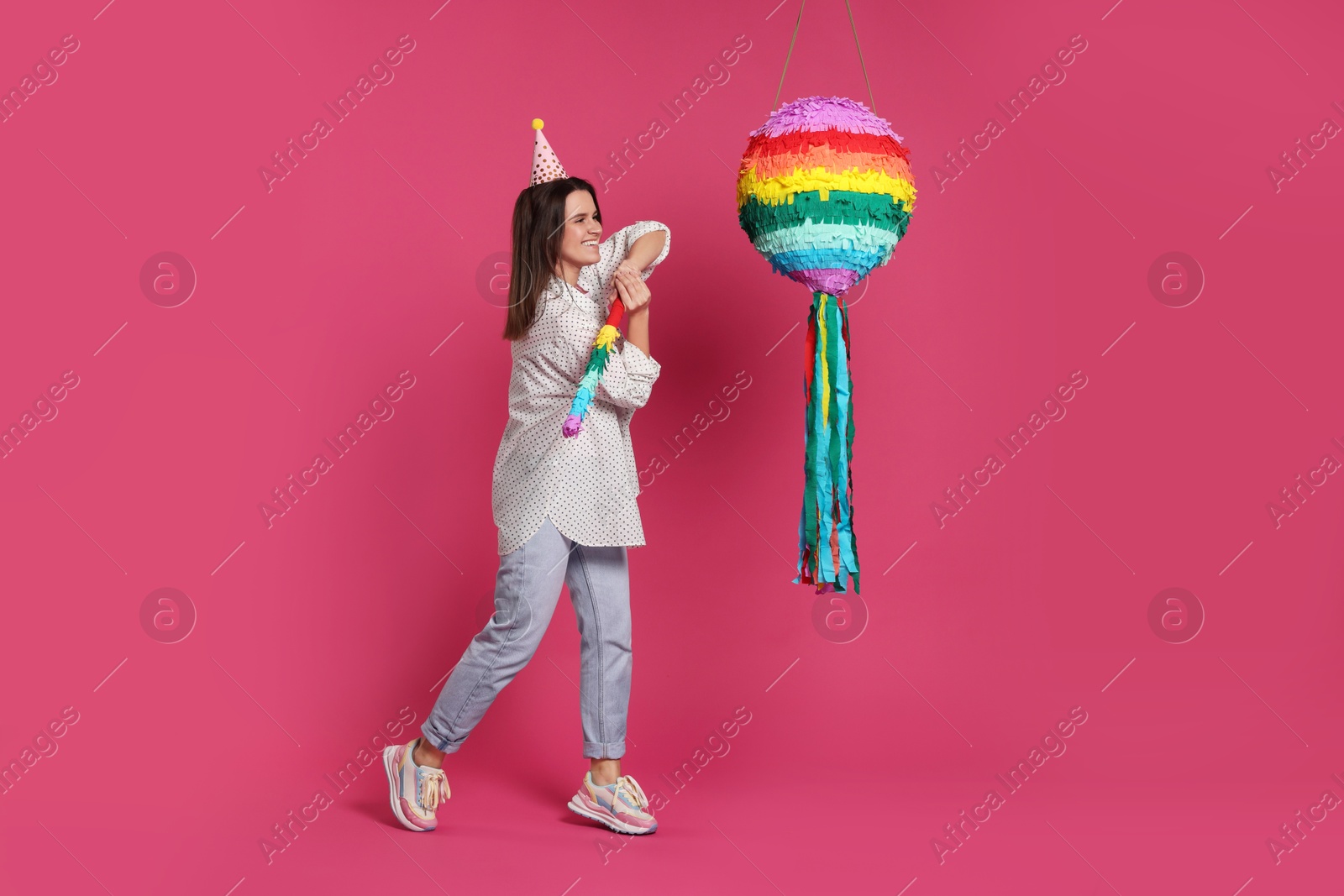 Photo of Happy woman hitting colorful pinata with stick on pink background