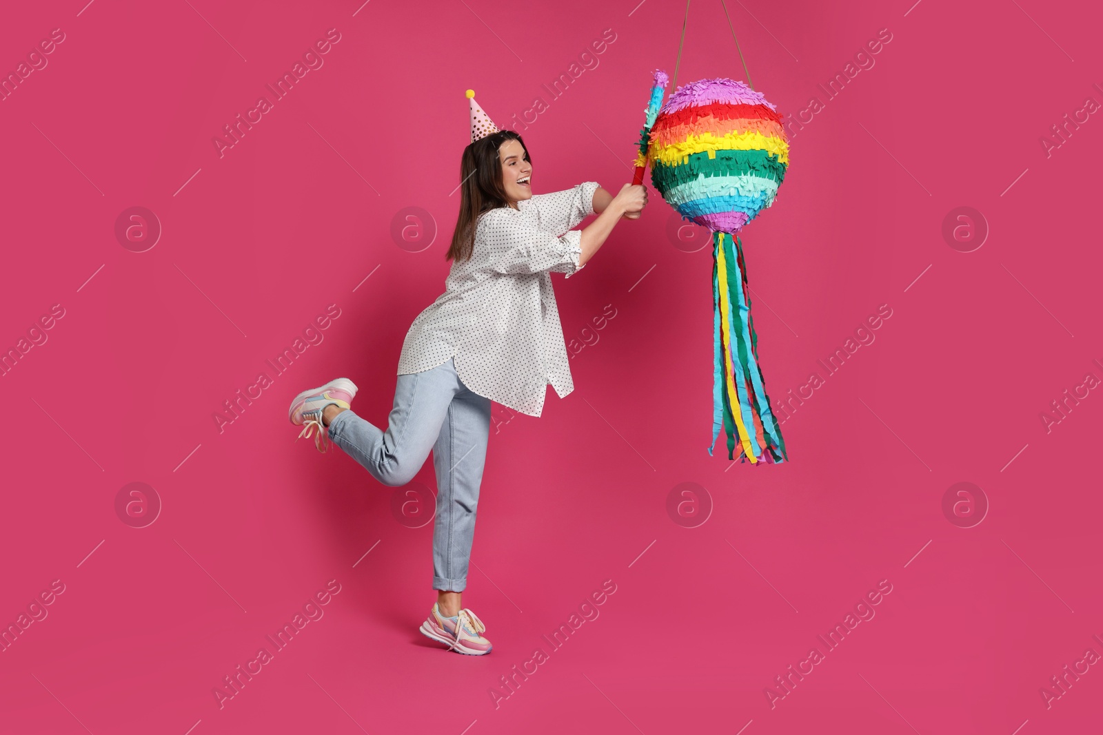 Photo of Happy woman hitting colorful pinata with stick on pink background