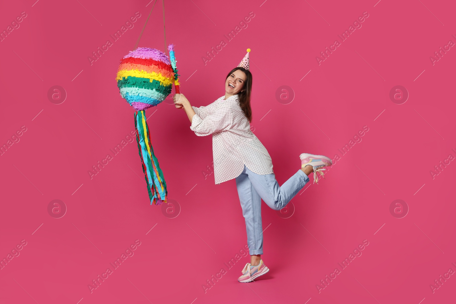 Photo of Happy woman hitting colorful pinata with stick on pink background