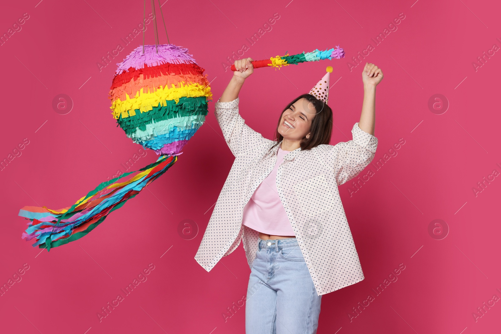 Photo of Happy woman hitting colorful pinata with stick on pink background