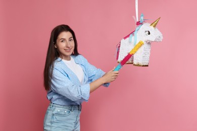 Happy woman hitting unicorn shaped pinata with stick on pink background