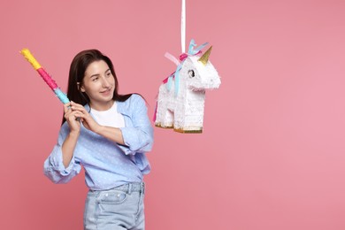 Photo of Happy woman hitting unicorn shaped pinata with stick on pink background, space for text