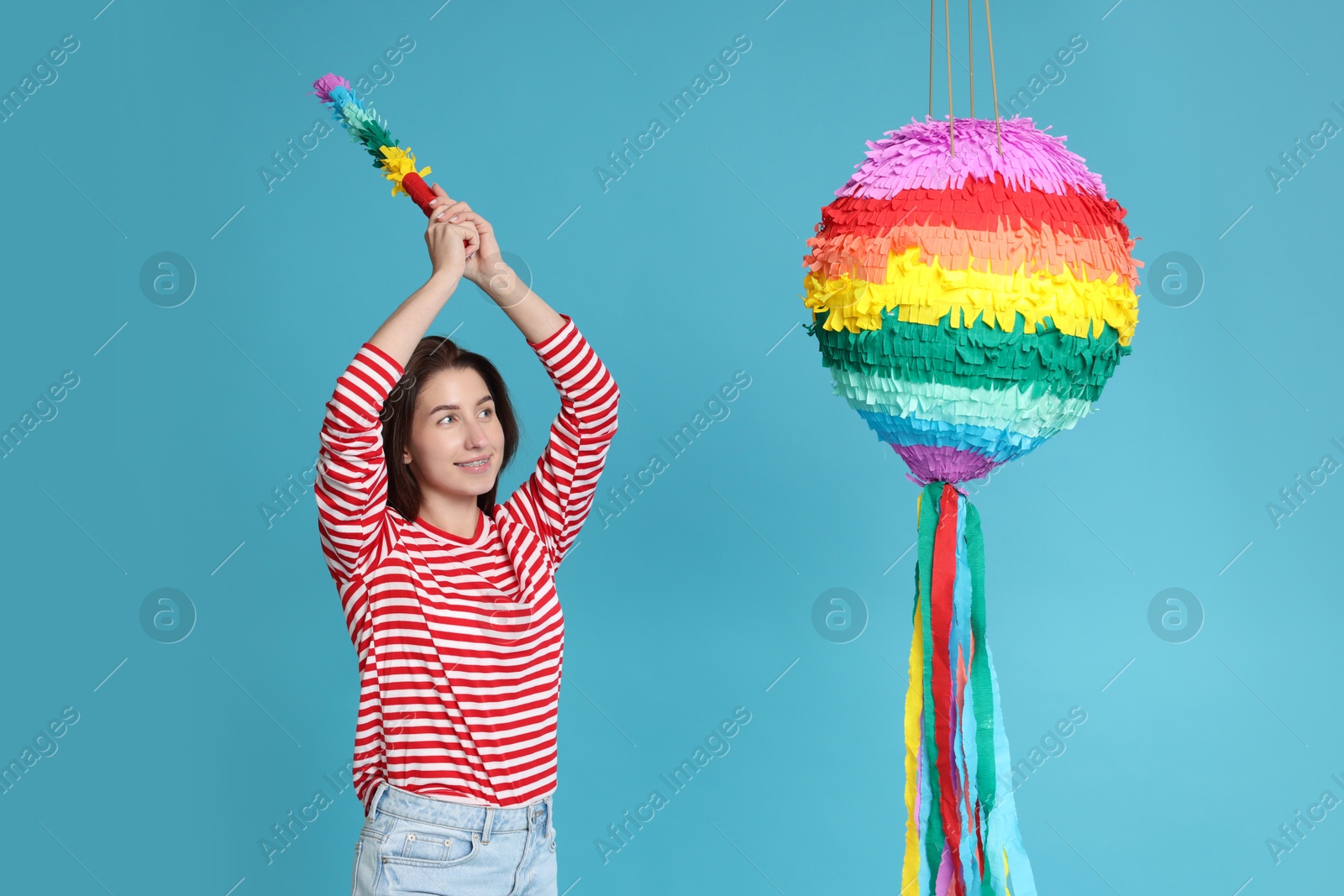 Photo of Happy woman hitting colorful pinata with stick on light blue background