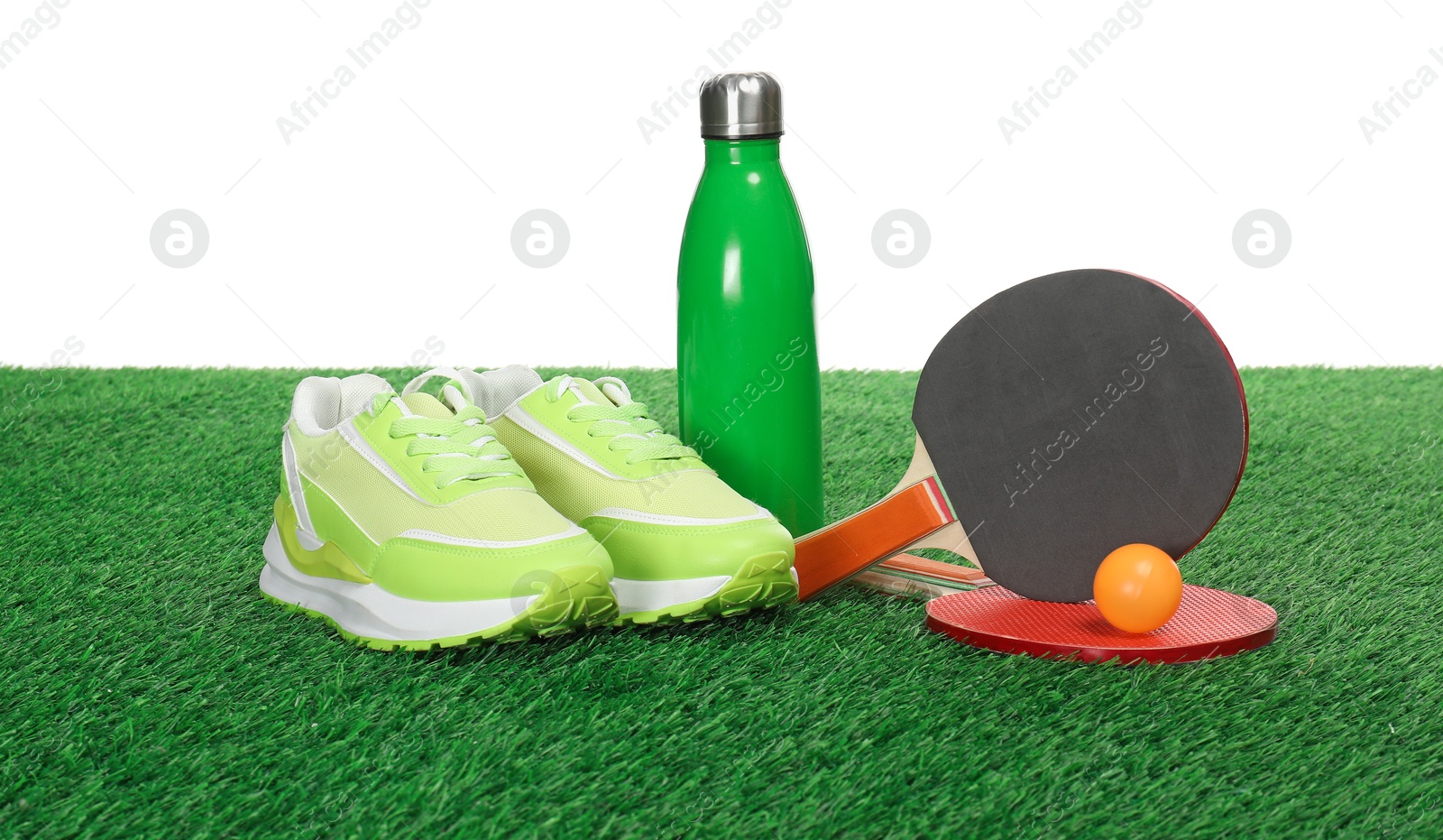 Photo of Ping pong rackets, ball, thermo bottle and sneakers on green grass against white background. Sport equipment