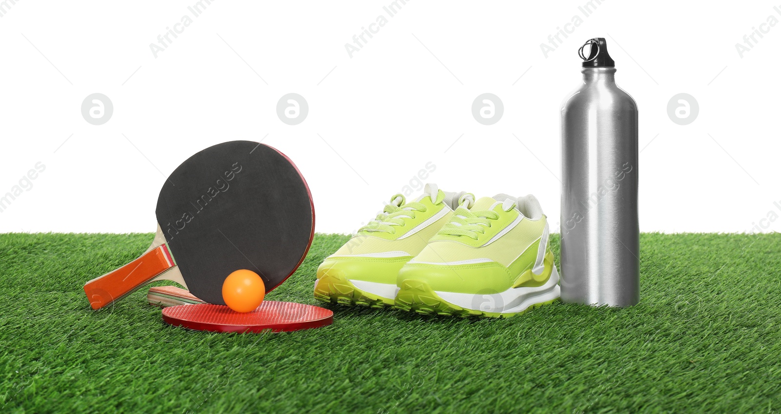 Photo of Ping pong rackets, ball, thermo bottle and sneakers on green grass against white background. Sport equipment