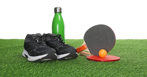 Photo of Ping pong rackets, ball, thermo bottle and sneakers on green grass against white background. Sport equipment