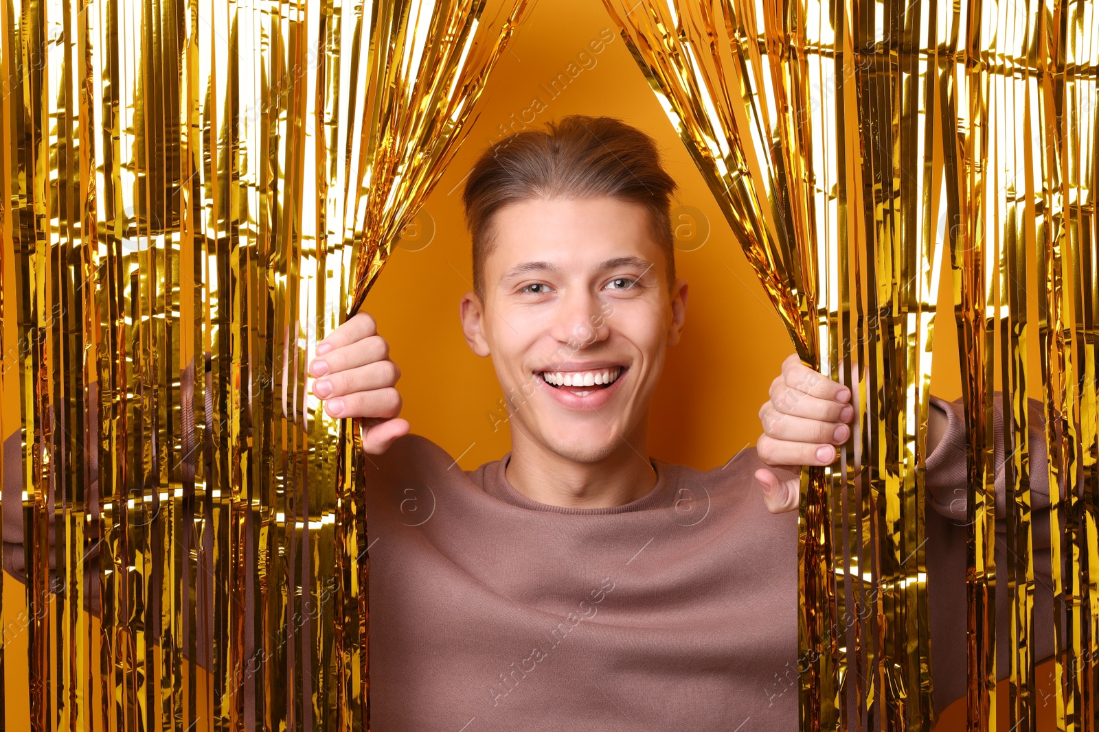 Photo of Happy young man looking out through golden foil curtain against orange background