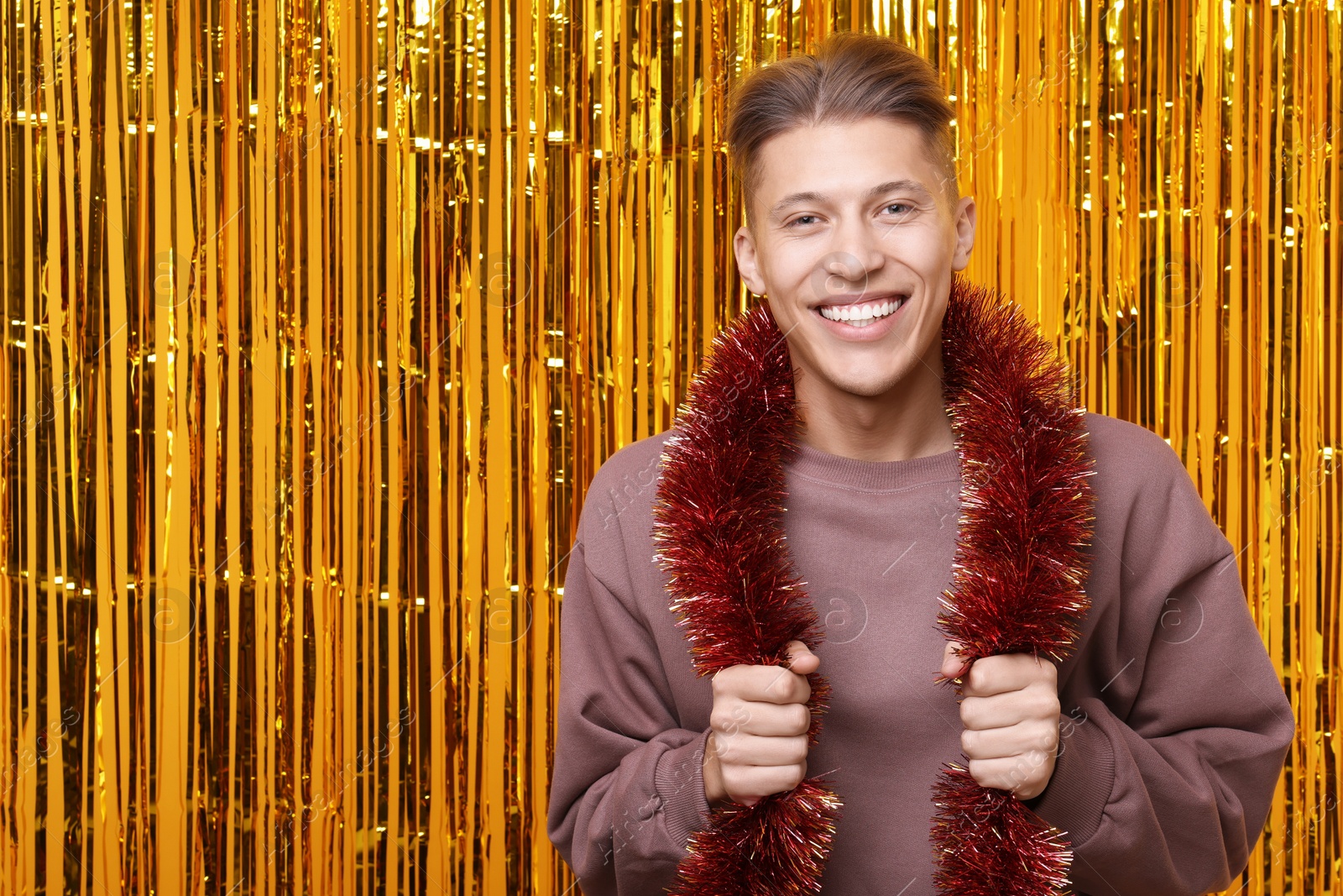 Photo of Happy young man with bright tinsel against golden foil curtain