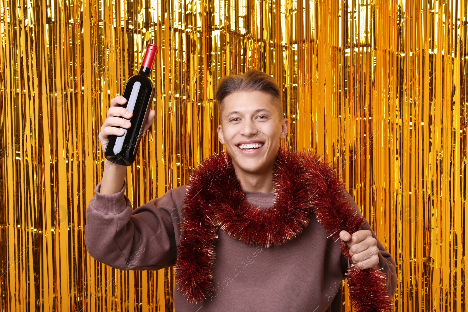 Photo of Happy young man with bright tinsel and bottle of wine against golden foil curtain